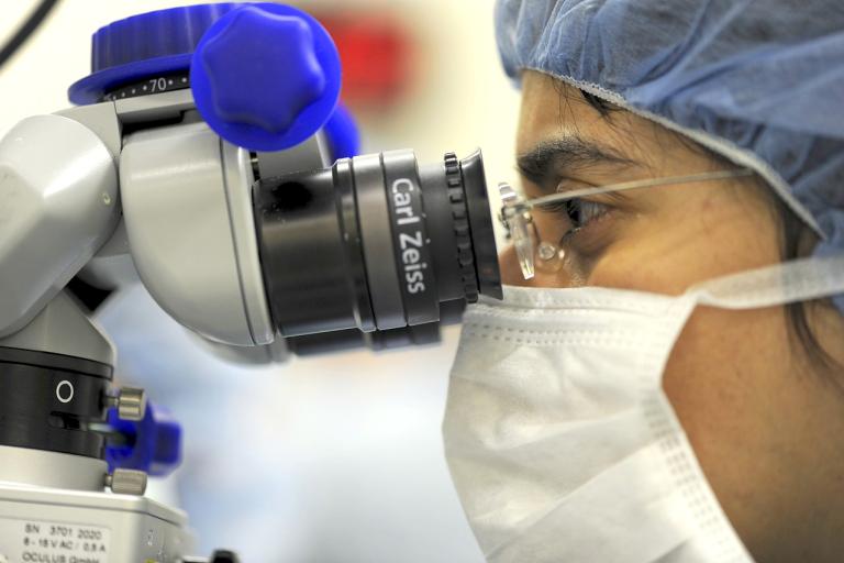 Female doctor, wearing a mask and reading glasses, looking into microscope.