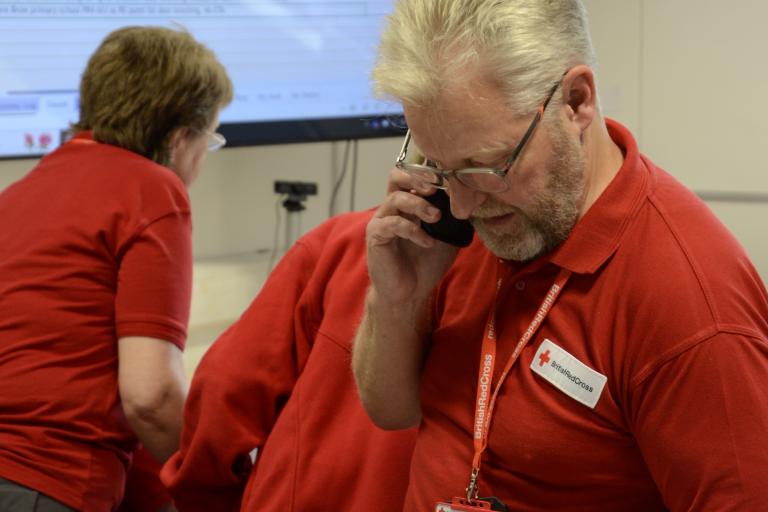 A male British Red Cross volunteer using a mobile phone and handheld radio in front of a large screen.
